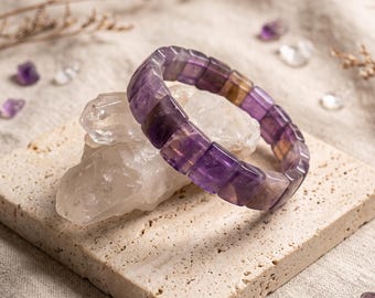 May include: A stretch bracelet composed of rectangular, polished beads made of purple and translucent amethyst stone. The beads feature varying shades of deep violet and light lavender, arranged in a continuous circular loop on a clear elastic cord.