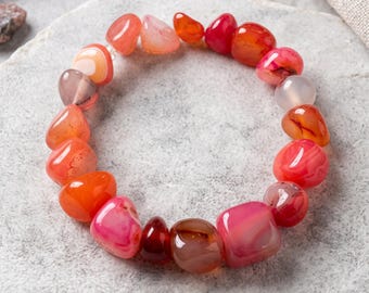 May include: A stretch bracelet composed of polished, irregular tumbled gemstone beads in various shades of red, orange, pink, and translucent white. The stones feature natural banding and smooth, rounded surfaces arranged in a circular loop.