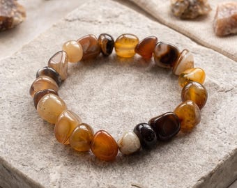 May include: A stretch bracelet composed of polished, tumbled gemstone beads in various shades of brown, amber, orange, and cream. The beads are irregular in shape and size, featuring natural banding and translucent qualities, strung together to form a circular loop.