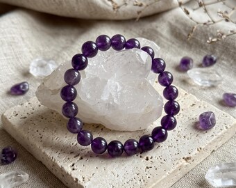 May include: A circular stretch bracelet composed of polished, deep purple amethyst beads. The bracelet is positioned around a large, translucent, jagged clear quartz crystal cluster, resting on a square, porous, beige stone tile.