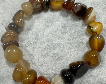 May include: A stretch bracelet composed of polished, tumbled agate stones in various shades of brown, amber, orange, and translucent cream. The smooth, rounded stones are strung together in a circular loop, showcasing natural colour variations.