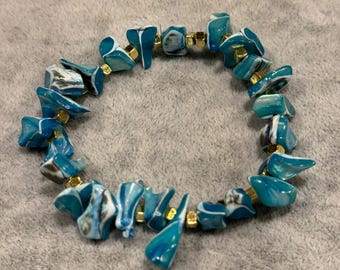 May include: A stretch bracelet featuring irregular, tumbled-style beads in shades of teal, blue, and white, interspersed with small, metallic gold-toned square spacer beads arranged in a circular pattern.