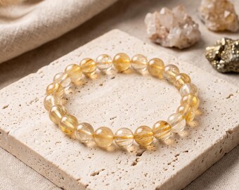 May include: A circular bracelet composed of polished, translucent golden rutilated quartz beads. Each bead features distinct, needle-like golden inclusions suspended within the clear stone, creating a shimmering, reflective appearance.