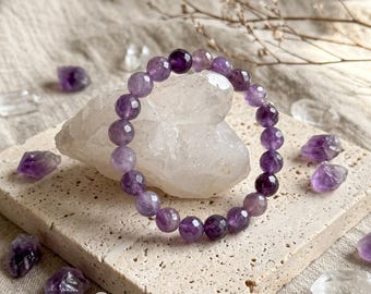 May include: A stretch bracelet composed of faceted, round purple amethyst beads. The beads exhibit varying shades of violet and translucent clarity, arranged in a circular pattern around a central, irregular clear quartz crystal.