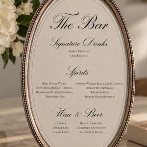 May include: An oval menu sign in a metallic bronze frame with ornate detailing at the top and bottom. The cream-colored insert features black text listing The Bar, Signature Drinks, Spirits, and Wine & Beer with various beverage names.