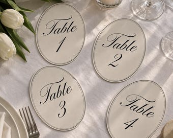 May include: Four oval-shaped off-white table number cards featuring black double-line borders and elegant black script text reading Table 1, Table 2, Table 3, and Table 4. Each card is printed on a smooth, matte-finish cardstock material.