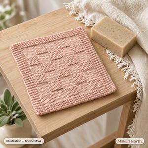 May include: A square, dusty pink knitted washcloth featuring a textured checkerboard pattern with a garter stitch border. The washcloth is positioned next to a rectangular bar of speckled beige soap and a cream-colored fringed textile.