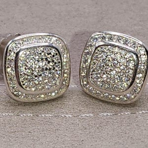 May include: A pair of silver-toned stud earrings featuring a square shape with rounded corners. Each earring is encrusted with numerous small, clear, sparkling stones covering the center and the border, creating a textured, reflective surface.