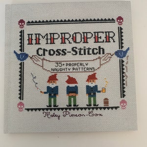 May include: A book titled "Improper Cross-Stitch: 35+ Properly Naughty Patterns" by Holly Pierson-Cox. The cover features a white background with a black border and three gnomes in blue and red clothing.