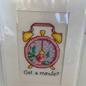 May include: Cross-stitch art of a pink and yellow alarm clock with a floral design inside the clock face. The text "Got a minute?" is stitched below the clock. The artwork is displayed in a white mat.