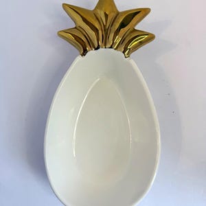 Puede incluir: Un plato de cerámica blanca con forma de piña y acabado liso y brillante. La parte superior del plato presenta un detalle de cerámica en color dorado metálico con forma de hojas puntiagudas de piña, creando un contraste decorativo al cuenco.