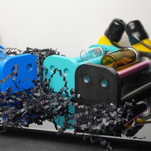May include: Two mechanical devices with blue and teal frames, black end plates, and attached yellow and black electrical components. Each unit features clear and amber glass vials held between the frames, connected by black power cords.