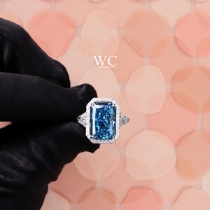 Può includere: Un anello con una grande pietra preziosa blu rettangolare a taglio radiante, montata su una fascia in metallo argentato e circondata da un alone di piccole pietre bianche. Il testo WC è visibile sopra l'anello su sfondo rosa.
