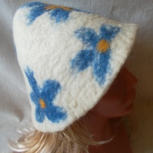 Peut inclure: Un chapeau en feutre blanc avec des motifs de fleurs bleues et jaunes. Le chapeau est porté sur une tête de mannequin avec des cheveux blonds.