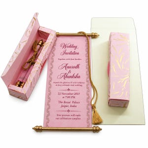 Peut inclure: Un faire-part de mariage en forme de parchemin rose avec des tiges métalliques dorées et un pompon doré. Le texte indique Wedding Invitation, Anurodh & Akanksha, 22 November 2027, The Royal Palace Jaipur, India. Inclut une boîte rose assortie.