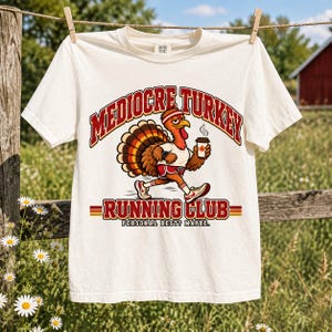 May include: An off-white short-sleeve t-shirt featuring a graphic of a turkey wearing a headband, t-shirt, and sneakers while holding a coffee cup. The text reads MEDIOCRE TURKEY RUNNING CLUB and PERSONAL BEST? MAYBE. The shirt has a crew neckline.