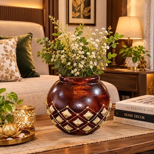 Puede incluir: Un jarrón redondo de madera marrón oscuro con un patrón decorativo de diamantes en tono dorado con pequeños tachones. El jarrón está lleno de un ramo de flores blancas y follaje verde, sobre una superficie de madera junto a libros apilados.
