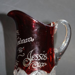 May include: A red glass pitcher with a clear glass handle. The pitcher has a cut glass design at the bottom and is etched with the words "From Tomans To Jessie From"