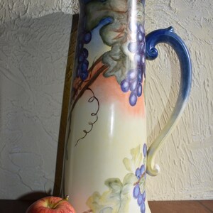 May include: A tall, cream-colored pitcher with a blue rim and handle, adorned with a hand-painted grape vine design. The pitcher has a curved handle and a wide base. A red apple rests beside the pitcher.