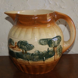 May include: A brown and cream colored ceramic pitcher with a green and brown painted design of trees and a house. The pitcher has a round body and a handle.