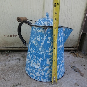 May include: A blue and white speckled enamel coffee pot with a metal handle, a wooden grip, and a lid. A yellow measuring tape is positioned vertically against the side of the pot, indicating a height of approximately 14 inches from base to top.