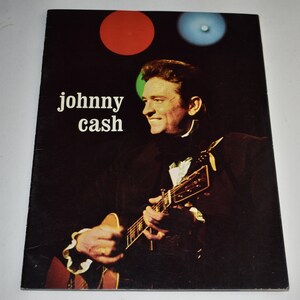 May include: A black and white photo of Johnny Cash playing a guitar on stage. The photo is from a concert and has red, green, and blue stage lights in the background. The text "johnny cash" is printed in white letters on a black background.