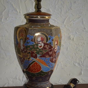 May include: A vintage Japanese-style table lamp with a ceramic base featuring a colorful hand-painted design of figures in gold, blue, red, and green. The lamp has a gold-colored metal base and a black cord.
