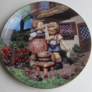 May include: A porcelain plate with a colorful illustration of two children standing near a small wooden bucket. The girl is wearing a pink and yellow dress and the boy is wearing blue overalls. The plate has a gold rim and is decorated with flowers and greenery.