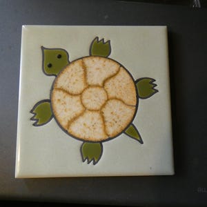 May include: A square ceramic tile featuring a stylized turtle illustration. The turtle has a light green head and four olive green flippers with dark outlines. The shell is a circular, textured beige piece with a cracked, crackle-glaze finish.