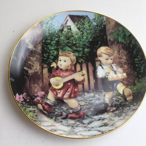 May include: A decorative porcelain plate featuring a colorful illustration of two children playing music in a whimsical outdoor setting. The girl plays a banjo while the boy plays a drum. The plate has a gold rim and a white background.