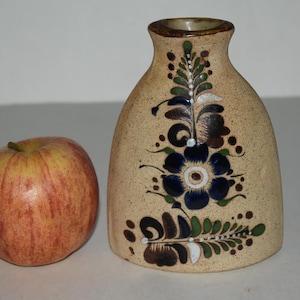 May include: A beige ceramic vase with a floral design in blue, green, and brown. The vase has a wide base and a narrow neck. An apple with red and yellow stripes is next to the vase.