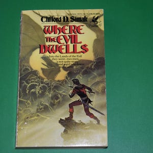 May include: A paperback book cover with a fantasy illustration of a man in armor standing on a rock with a sword in his hand. The book title is "Where the Evil Dwells" by Clifford D. Simak.