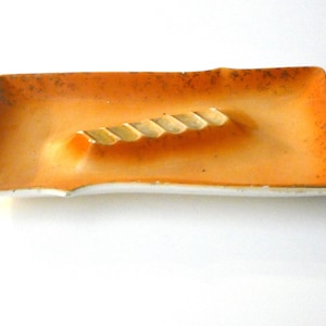 May include: Rectangular ceramic ashtray with a warm orange and brown gradient. The ashtray features a central rest with a rope-like design. The edges are slightly irregular, and the base is white.