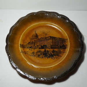 May include: A decorative plate with a brown and gold color scheme. The plate features a detailed illustration of a building with a dome and columns, likely a government building. The plate has a scalloped edge and text that reads "The Capitol, Washington, D.C."