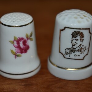 May include: Two white ceramic thimbles. One thimble has a pink rose design. The other thimble has a black and white image of Elvis Presley with the text "Elvis Presley" and "CEPE".