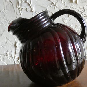 May include: A dark red glass pitcher with a ribbed design. The pitcher has a rounded body, a curved handle, and a spout for pouring. The pitcher is approximately 9 inches tall.