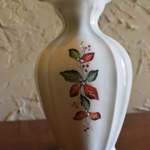 May include: White ceramic vase with a ruffled top and a floral design featuring red and green leaves. The vase has a textured, ribbed surface.