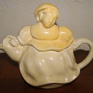 May include: A yellow ceramic teapot shaped like a woman wearing a dress and bonnet.