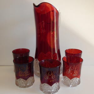 May include: A ruby red glass pitcher and matching glasses set. The pitcher is tall with a flared top, and the glasses are short with a textured base. Gold lettering and the year 1907 are visible on the glasses.