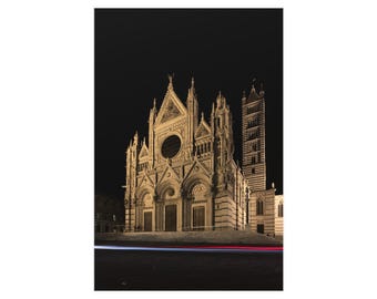 Op de afbeelding: Een nachtelijk uitzicht op de kathedraal van Siena, een groot stenen gebouw met complexe gotische architectuur, een centraal roosvenster en een hoge gestreepte klokkentoren, verlicht tegen een donkere lucht met een lichtspoor op de voorgrond.