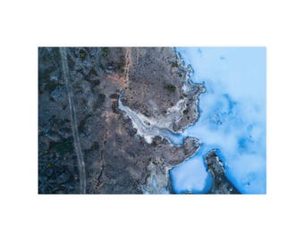 Op de afbeelding: Een luchtfoto van een landschap met donkere, ruige vulkanische rotsformaties aan de linkerkant, contrasterend met een levendig, melkachtig lichtblauw geothermisch zwembad aan de rechterkant, gescheiden door een grillige kustlijn.