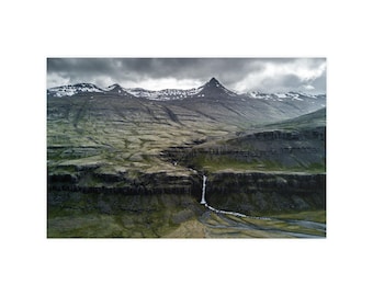 Op de afbeelding: Een landschapsfoto van een ruig gebergte met besneeuwde toppen onder een bewolkte lucht. Een smalle waterval stroomt langs een donkere, gelaagde rotswand naar beneden in een vallei met groene vegetatie en een kronkelende beek.