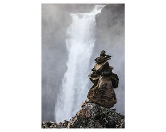 Op de afbeelding: Een stapel van vijf onregelmatige, donkerbruine stenen die verticaal in evenwicht zijn gehouden op een grote, bemoste rotsformatie. Op de achtergrond stroomt een hoge, krachtige waterval langs een steile, mistige klif, wat een zacht, wazig wit watereffect creëert.