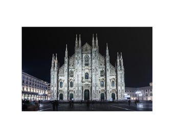 Op de afbeelding: De kathedraal van de Duomo di Milano staat 's nachts verlicht en toont zijn complexe gotische architectuur, talloze torenspitsen en gedetailleerde stenen gevel. Het bouwwerk is vastgelegd tegen een donkere lucht en benadrukt het ontwerp.
