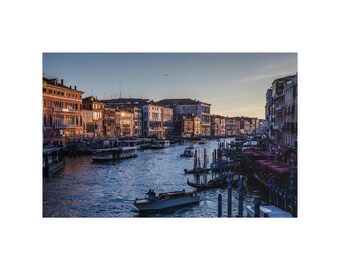 Op de afbeelding: Een panoramisch uitzicht op het Canal Grande in Venetië bij zonsondergang, met historische gebouwen van meerdere verdiepingen met warme oranje en bruine gevels langs het water, talloze boten en gondels, en een heldere avondlucht.
