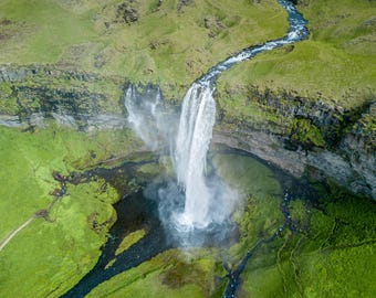 Op de afbeelding: Een luchtfoto van een hoge waterval die over een rotsachtige klif in een donkere poel stroomt. Het omliggende landschap bestaat uit felgroene grashellingen, met mos begroeide rotsformaties en een kronkelende rivier.