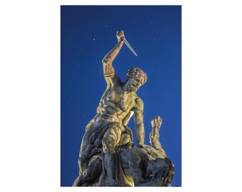 Op de afbeelding: Een stenen sculptuur die een gespierde figuur voorstelt die boven een liggende figuur staat, waarbij de staande figuur een scherp, puntig mes hoog boven het hoofd houdt tegen een donkerblauwe sterrenhemel.
