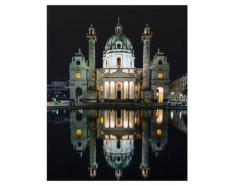 Op de afbeelding: De Karlskirche in Wenen is 's nachts verlicht, met een grote centrale koepel, twee hoge zuilen en een symmetrische gevel. Het gebouw wordt weerspiegeld in het donkere water op de voorgrond, wat een helder spiegelbeeld creëert.