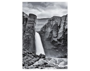 Op de afbeelding: Een zwart-wit landschapsfoto van een waterval die naar beneden stroomt tussen twee hoge, grillige, donkere rotswanden. Het water stroomt in een rotsachtig bekken aan de voet, terwijl een bewolkte lucht boven het bergachtige terrein hangt.
