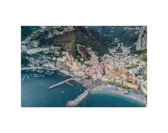 Op de afbeelding: Een luchtfoto van het kuststadje Positano in Italië, met veelkleurige gebouwen genesteld in de steile kliffen boven de diepblauwe Tyrreense Zee, inclusief een zandstrand en twee stenen pieren die in het water uitsteken.